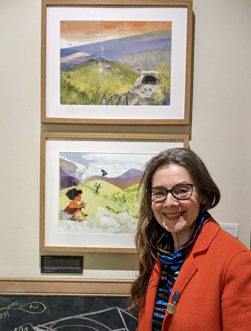 Bridget standing in front of 2 framed illustrations of landscapes from her book Mo's Best Friend, A Stone Age Story